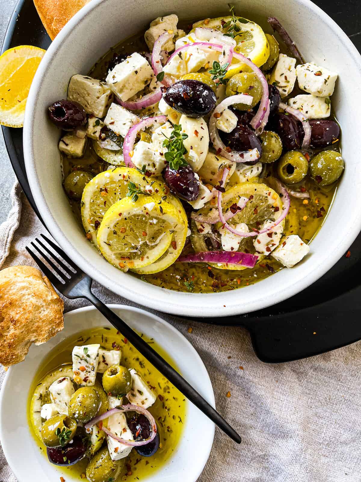 bowl of marinated olives and feta alongside crusty bread for serving and a small side plate of the mixture.