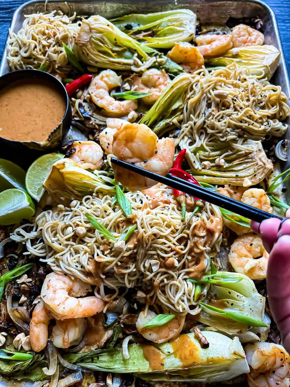 Asian shrimp held by a chopstick with sheet pan of noodles, shrimp, peanut sauce and garnishes in the background