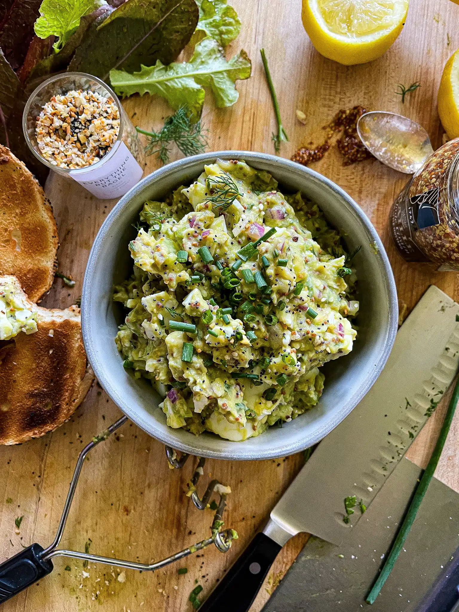 avocado egg salad on a toasted everything bagel above a cutting board with the recipe ingredients and equipment