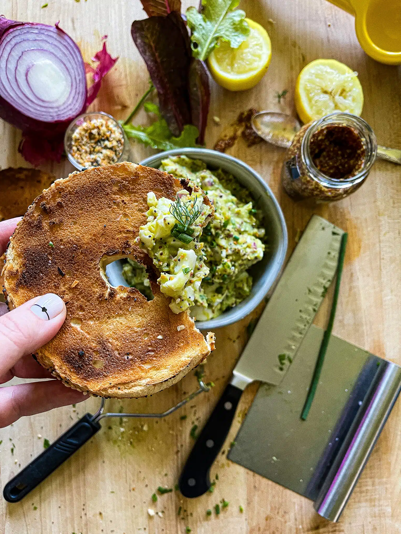 avocado egg salad on a toasted everything bagel above a cutting board with the recipe ingredients and equipment