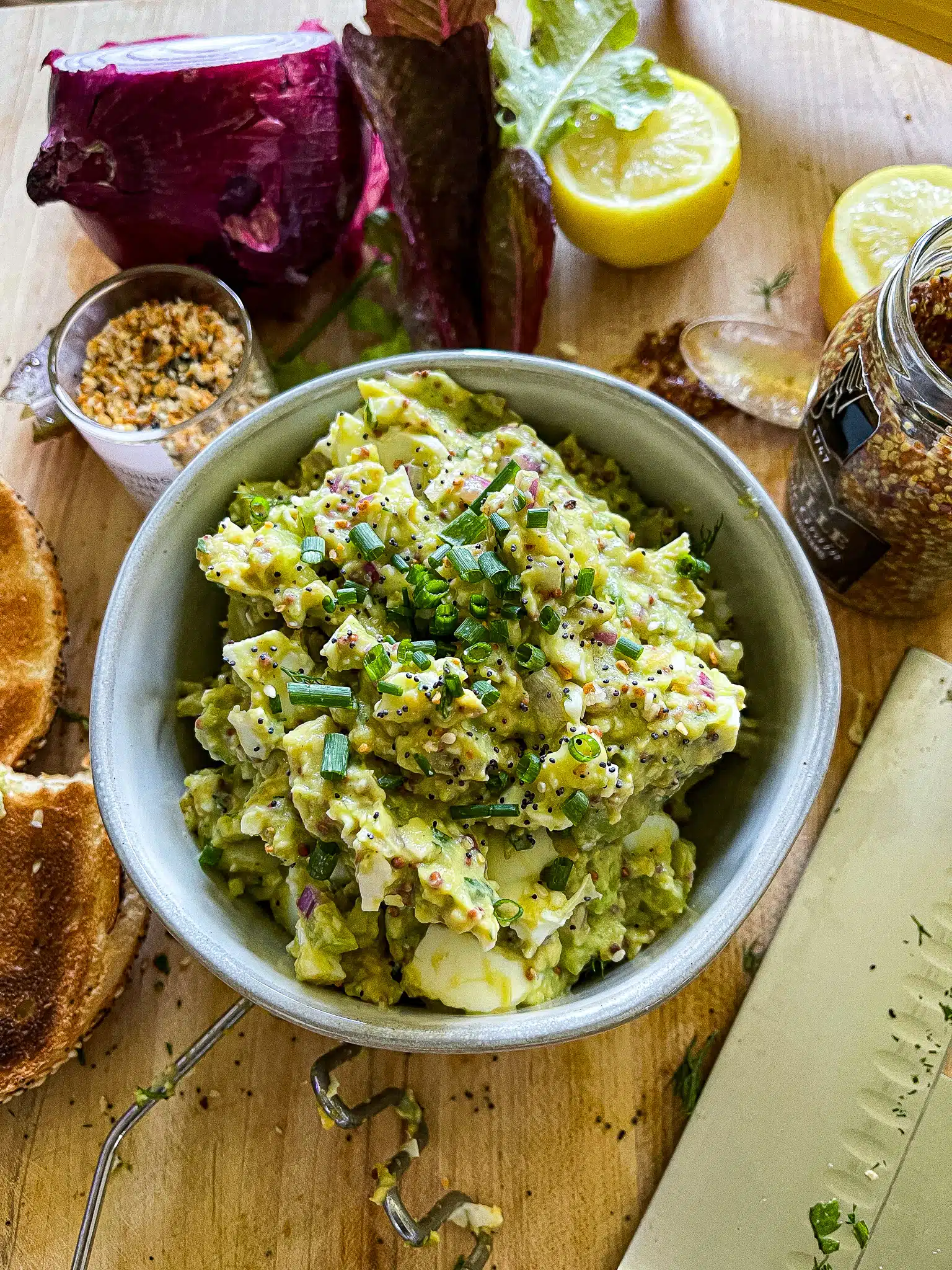 avocado egg salad on a toasted everything bagel above a cutting board with the recipe ingredients and equipment