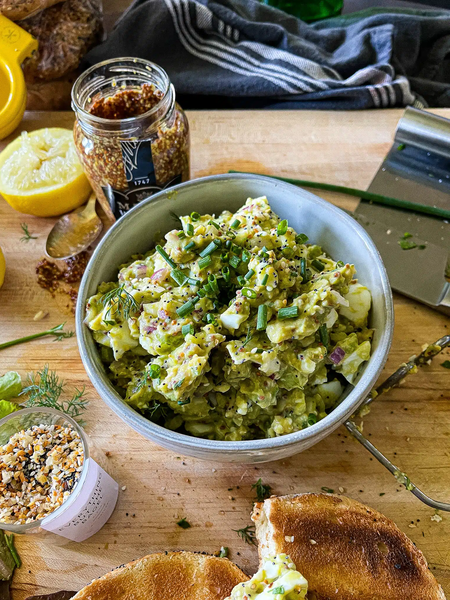 avocado egg salad on a toasted everything bagel above a cutting board with the recipe ingredients and equipment