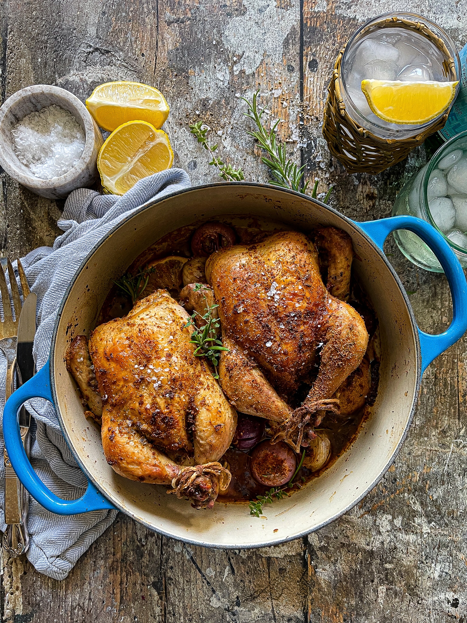 This is a picture of a small blue dutch oven with two roasted cornish hens. There is a container of flaky sea salt and fresh sliced lemons to the side. 