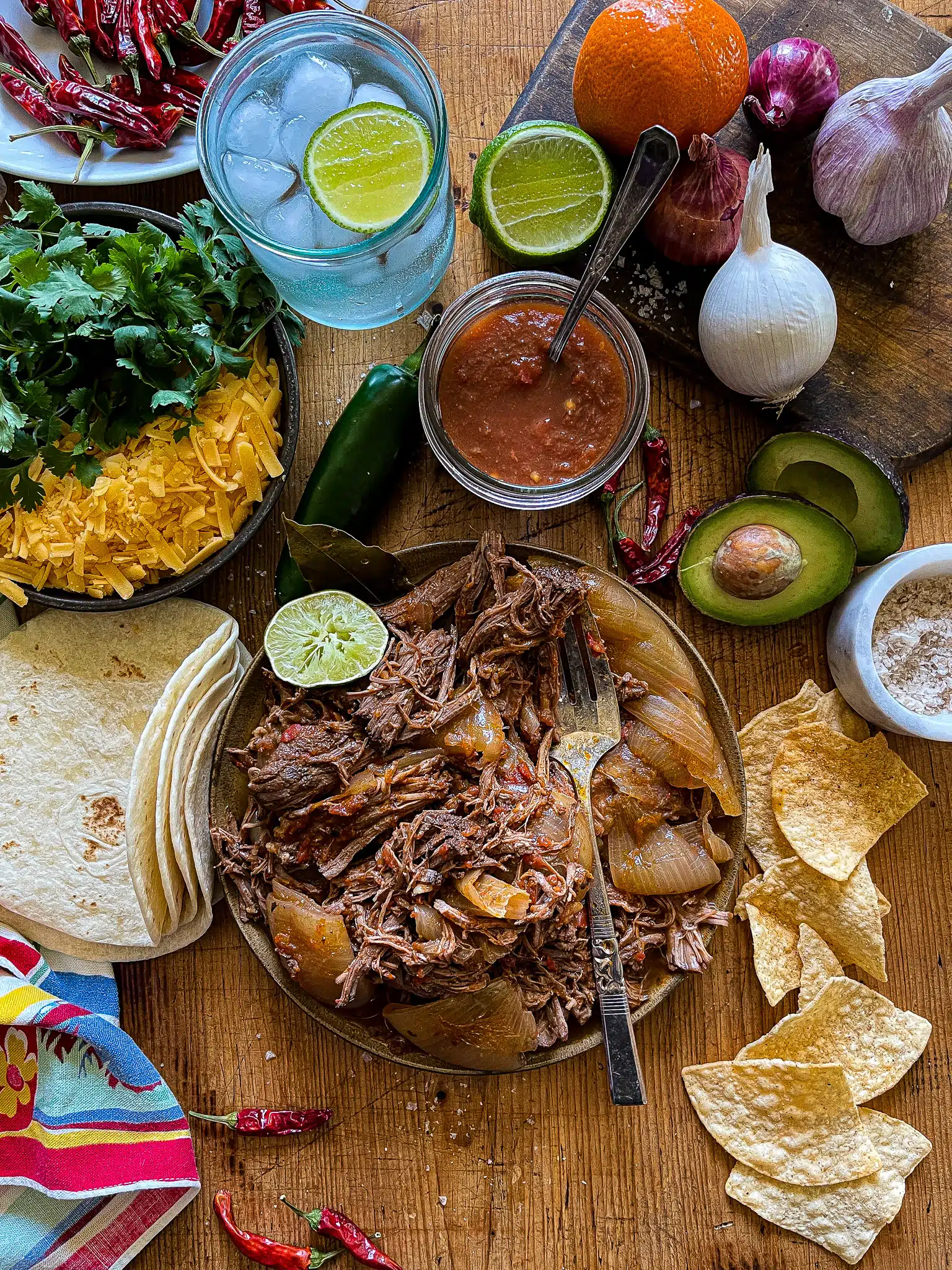 This is a picture of slow cooker Mexican Beef with tortillas, chips, avocado halves, salsa, shredded cheese, and lime wedges on the side.