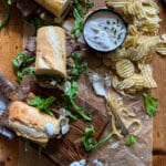 This is a picture of a steak sandwich cut into portions with a side of potato chips and horseradish sauce.