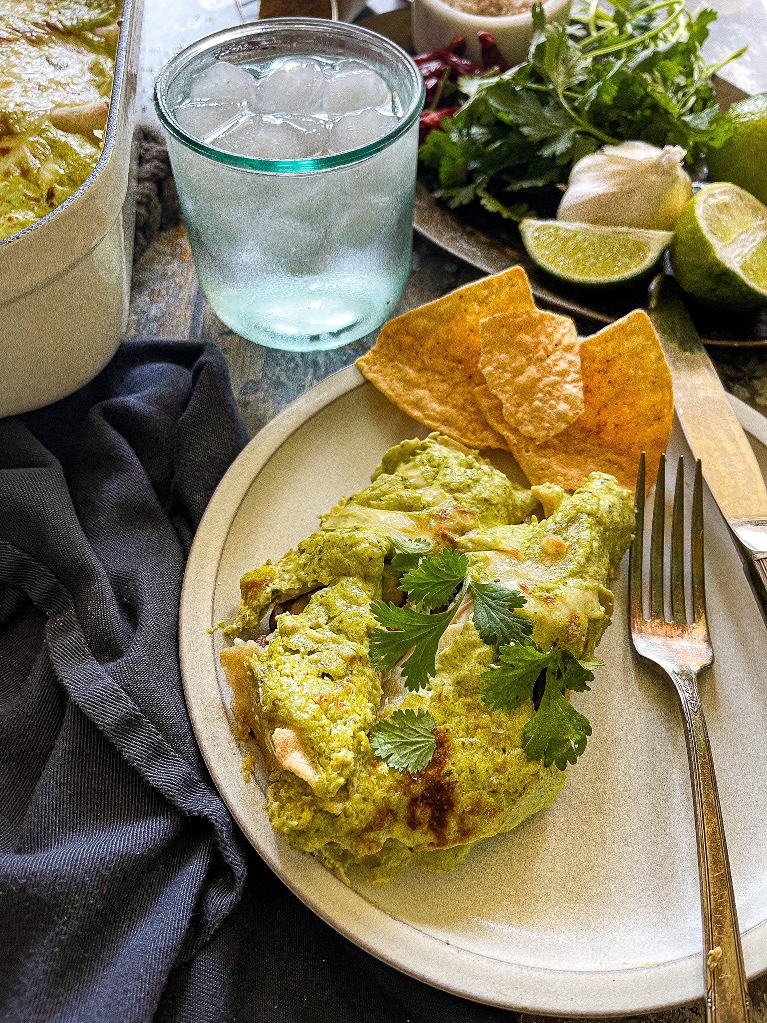 This is a plate of chicken enchiladas with a poblano cream sauce. There is a glass of ice water and a plate of garnishes such as cilantro and extra lime on the side. 