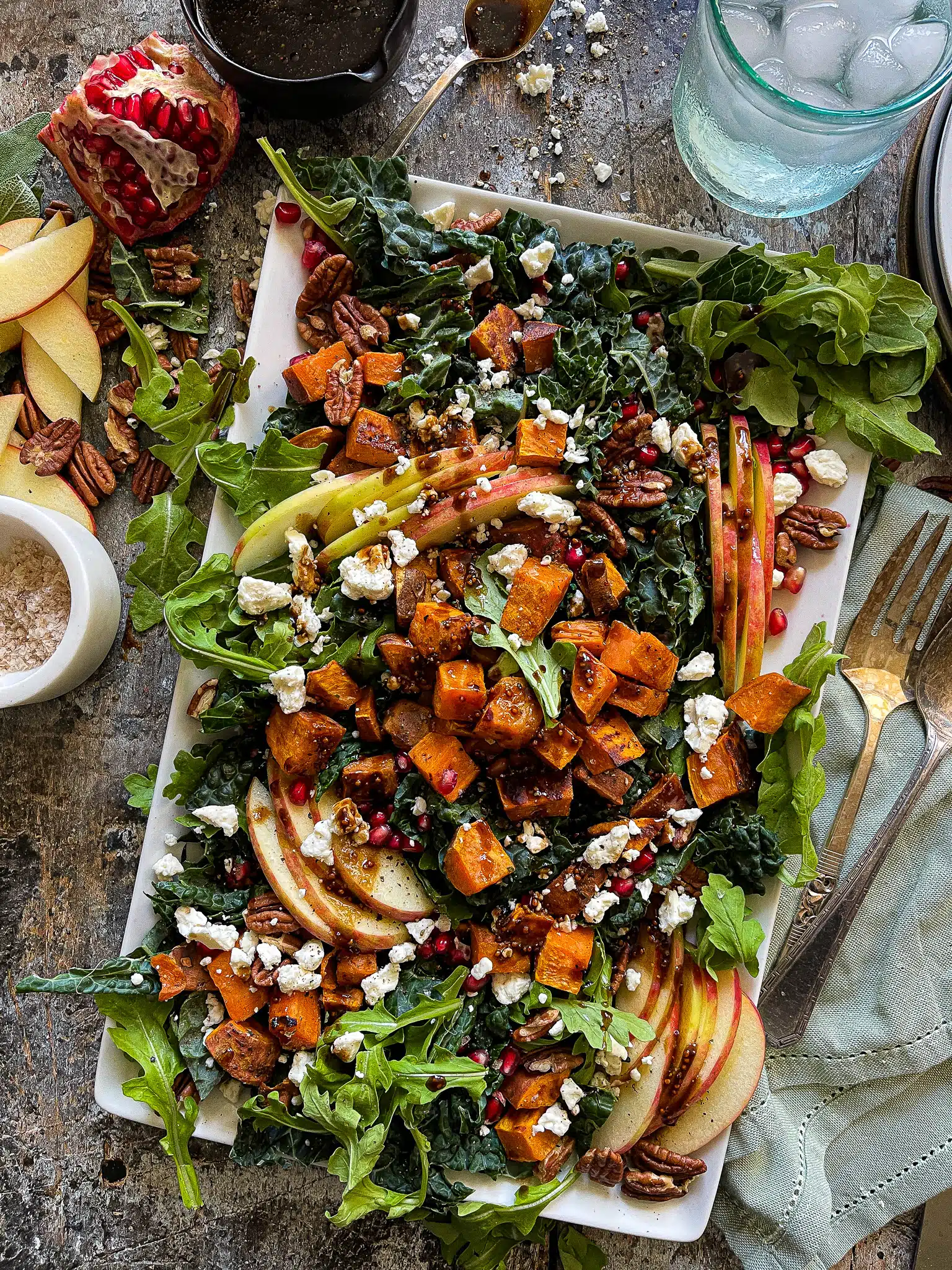 This is a picture of a Fall Harvest Salad on a large serving platter flanked by pomegranate, apple slices, and balsamic vinaigrette.