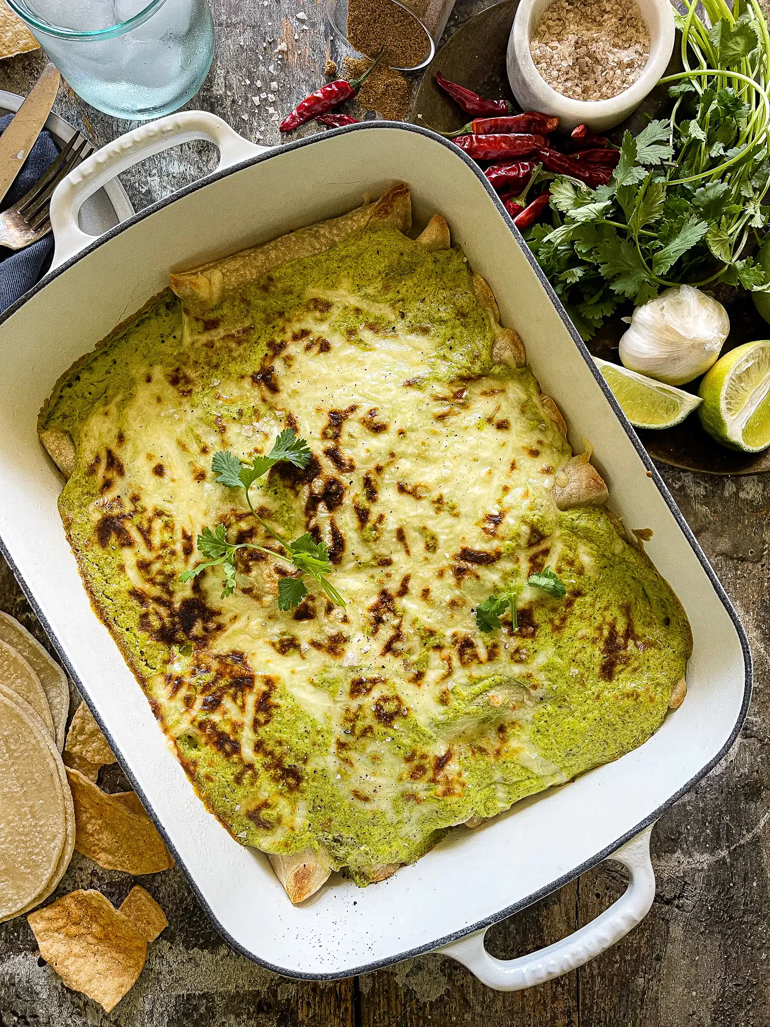 This is a 9x13 baking dish with Poblano Cream Chicken Enchiladas. There is a plate to the side of garnishes with extra cilantro and lime wedges.