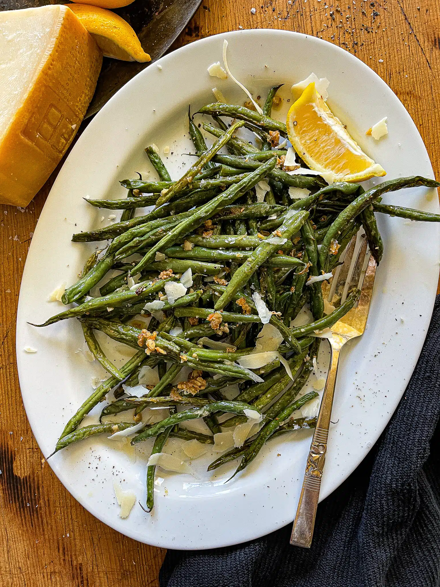 This is a serving platter with Roasted Green Beans and Garlic. There is a wedge of lemon and a vintage serving fork to the side.