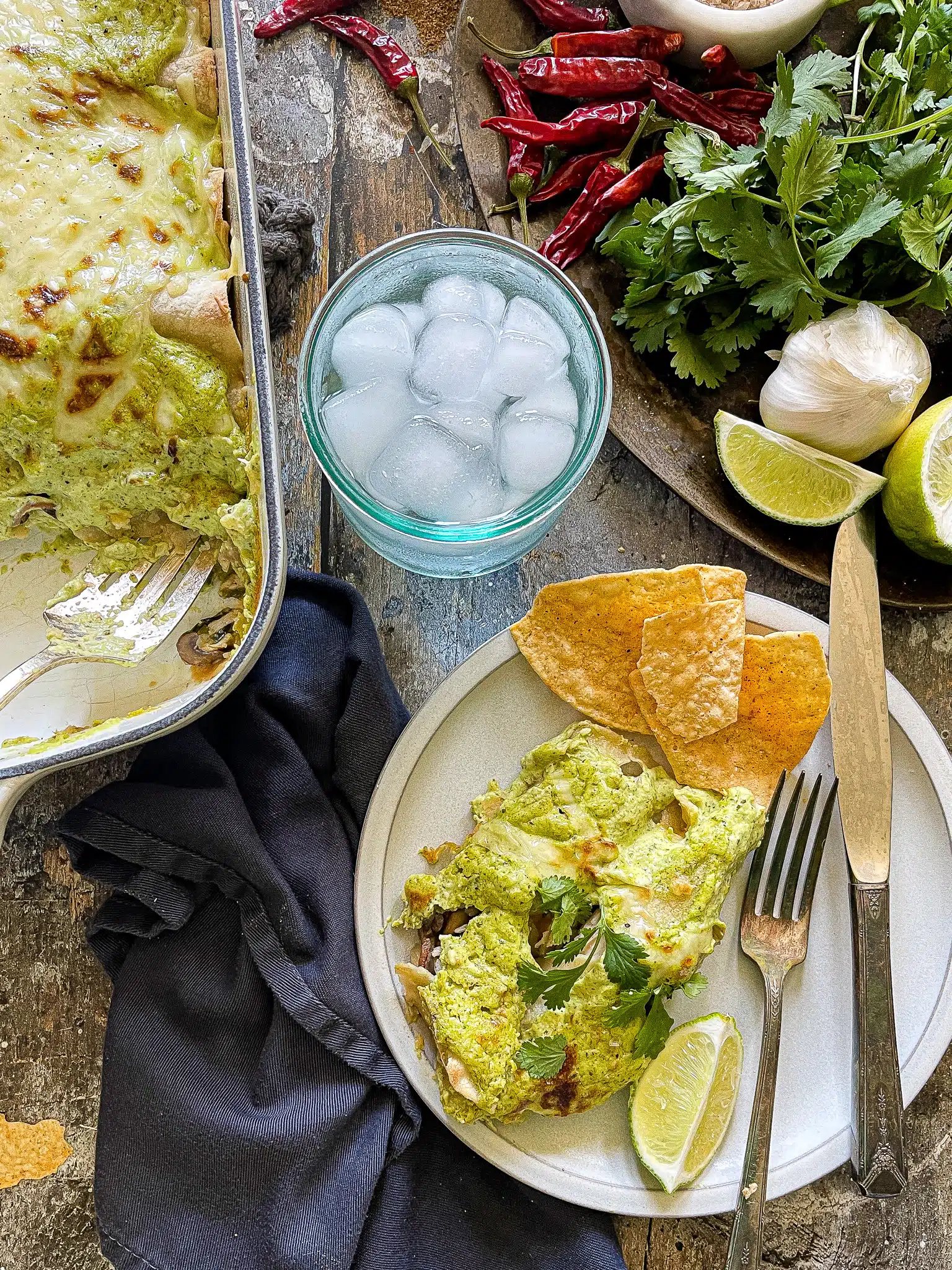 This is a plate of chicken enchiladas with a poblano cream sauce. There is a glass of ice water and a plate of garnishes such as cilantro and extra lime on the side. 