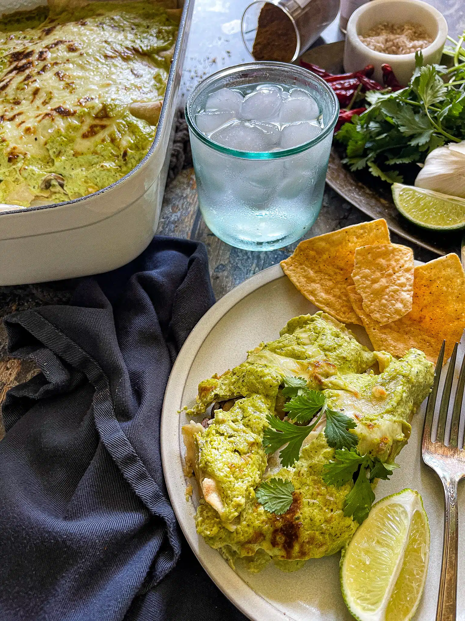 This is a plate of chicken enchiladas with a poblano cream sauce. There is a glass of ice water and a plate of garnishes such as cilantro and extra lime on the side. 