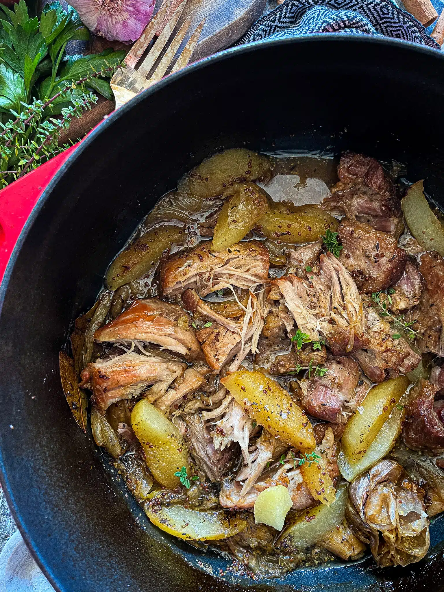 This is a picture of braised pork shoulder in a dutch oven with the cooked apple slices. There is fresh sage, thyme, and parsley on the side.