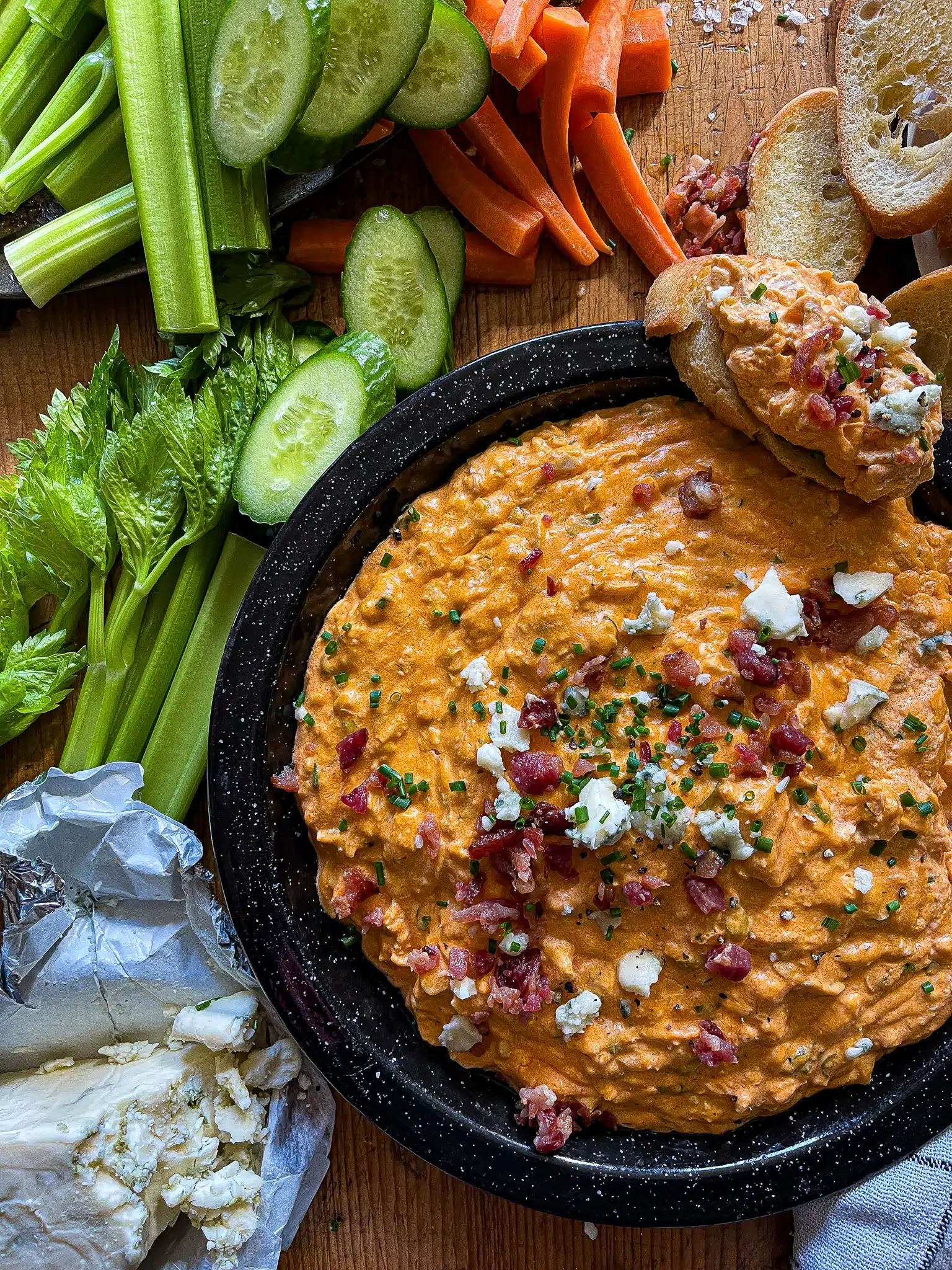 This is a picture of Slow Cooker Buffalo Chicken Dip topped with blue cheese crumbles and bacon bits. It's surrounded by sliced carrots, celery, and cucumbers along with a pile of crostini.