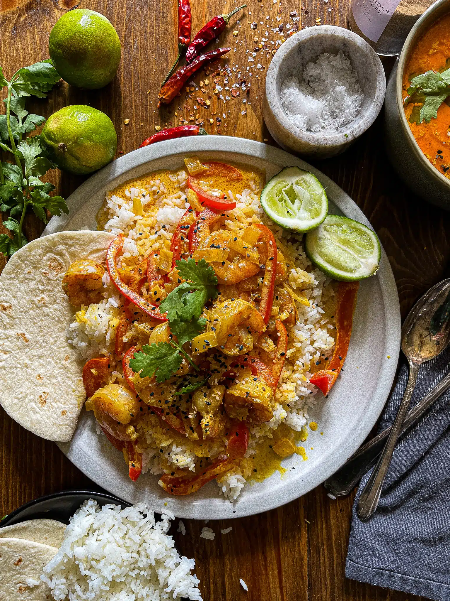 This is a picture of Shrimp Coconut Curry with red bell peppers spooned over white rice. Garnished with black and white sesame seeds, cilantro leaves, and lime.