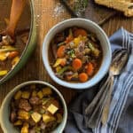 This is a picture of bowls of vegetable beef soup with chunks of chuck roast, carrots, green beans, potato, and mushrooms in a deep brown broth. There are spoons sitting on top of cloth napkins to the side.