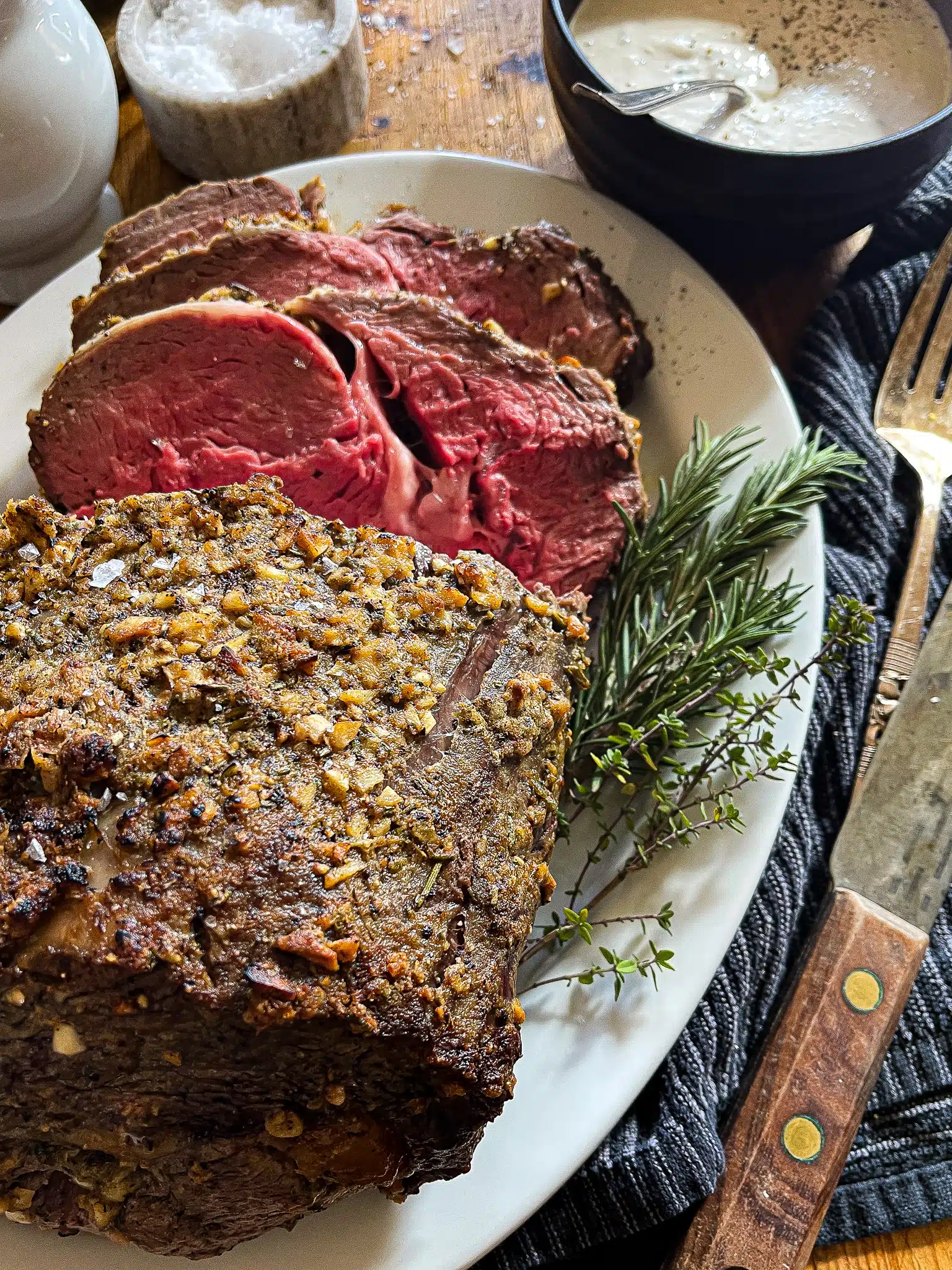 this is a picture of a roasted prime rib on a white serving platter with fresh rosemary and thyme sprigs as a garnish