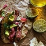 this is a picture of a steak taco in small flour tortillas with a creamy sauce, pickled onions, avocado and fresh cilantro leaves on a parchment paper background