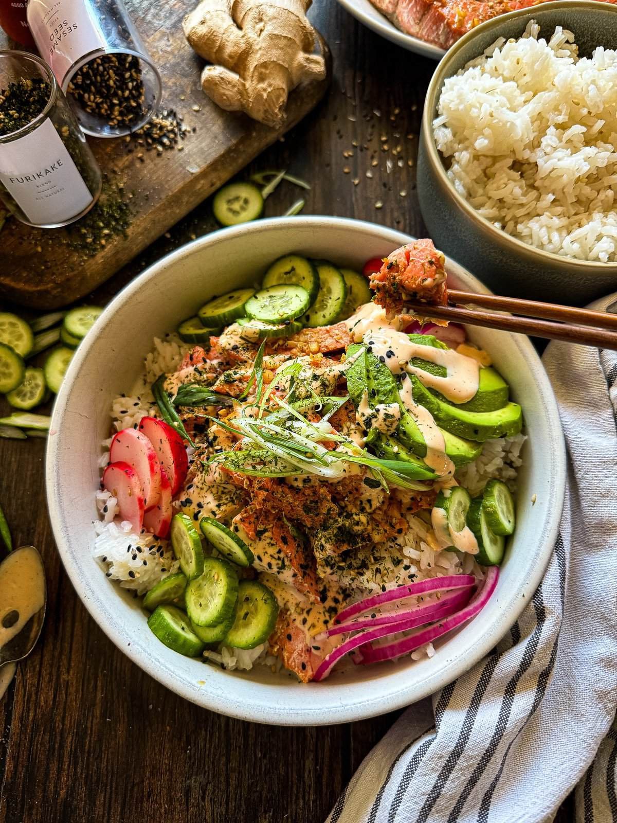 this is a picture of a sushi bowls with pickled onions and radish, salmon, avocado and a green onion garnish with a side of spicy sriracho mayo
