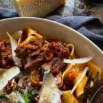 picture of a white serving bowl of slow cooker beef ragu topped with parmesan cheese shavings and fresh parsley garnish. sitting on a wooden surface with a navy napkin.