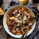 picture of a white serving bowl of slow cooker beef ragu topped with parmesan cheese shavings and fresh parsley garnish. sitting on a wooden surface with a navy napkin.