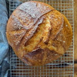 Loaf of gluten-free no-knead bread resting on a wire baking rack on a wooden surface next to a dish towel