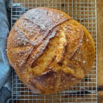 Loaf of gluten-free no-knead bread resting on a wire baking rack on a wooden surface next to a dish towel