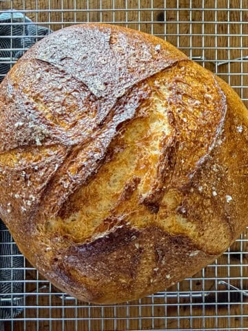 Loaf of gluten-free no-knead bread resting on a wire baking rack on a wooden surface next to a dish towel